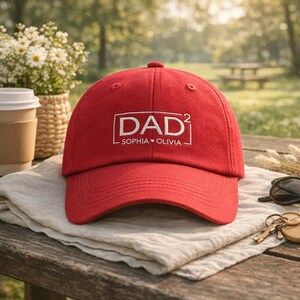 May include: A red baseball cap with the word "DAD²" embroidered in white, along with "SOPHIA • OLIVIA" underneath. The cap is sitting on a light-colored cloth on a wooden table. Sunglasses and keys are also on the table.