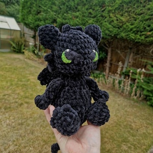 May include: A handmade black crochet dragon plush toy with bright green eyes. The plush has small wings and is made of a soft, textured yarn. The dragon is a popular character from the "How to Train Your Dragon" movie series.