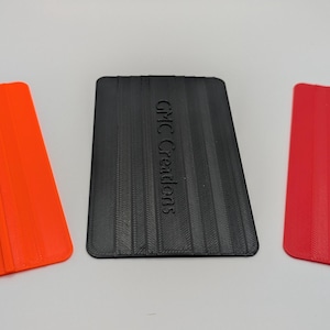 Puede incluir: Tres rasquetas de plástico en naranja, negro y rojo. Las rasquetas negras y rojas tienen el texto "GMC Creations" en relieve. Las rasquetas tienen forma rectangular con un borde biselado.