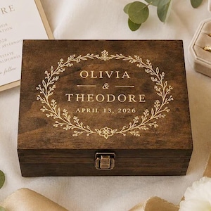 May include: A rectangular dark brown wooden box with a metal latch. The lid features a gold-colored engraved floral wreath surrounding the text OLIVIA & THEODORE and the date APRIL 13, 2026, centered on the top surface of the wooden container.