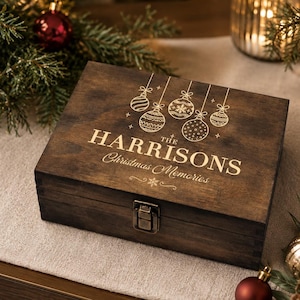 May include: A rectangular dark brown wooden keepsake box with a metal latch. The lid features gold-colored engraved text that reads THE HARRISONS Christmas Memories, accompanied by a graphic illustration of five hanging Christmas ornaments.