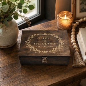 May include: A rectangular dark brown wooden box with a metal latch. The lid features a gold-colored engraved floral wreath surrounding the text OLIVIA & THEODORE and the date APRIL 13, 2026, centered on the top surface of the wooden container.