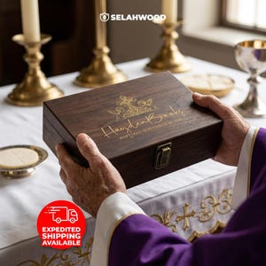 May include: A dark brown wooden box with a gold clasp and engraved text. The box features a cross and angel design, with the name "Hayden Brooks" and the date "Baptized September 26, 2026" in gold lettering. The box is held by a person wearing a purple robe.