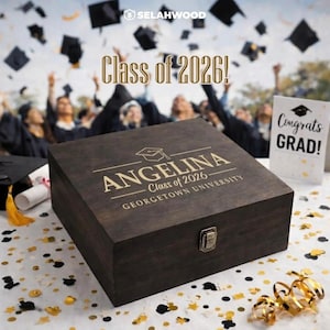 May include: A dark wooden graduation box with the name "ANGELINA" and "Class of 2026" engraved on the lid. The box is closed with a metal clasp. The background shows people throwing graduation caps in the air.