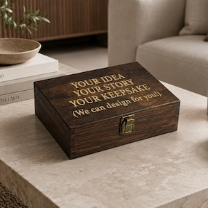 May include: A rectangular dark brown wooden box with a hinged lid and a brass-colored metal latch. The top of the lid features gold-colored engraved text that reads: YOUR IDEA YOUR STORY YOUR KEEPSAKE (We can design for you!).