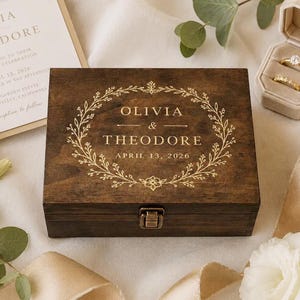 May include: A rectangular dark brown wooden box with a metal latch. The lid features a gold-colored engraved floral wreath surrounding the text OLIVIA & THEODORE and the date APRIL 13, 2026, centered on the top surface of the wooden container.