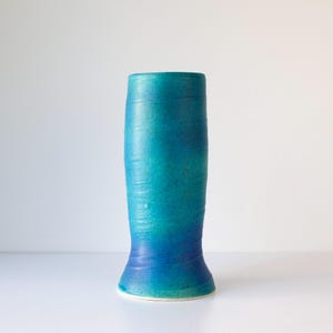 May include: A tall, handcrafted ceramic vase with a gradient of teal and blue hues. The vase has a cylindrical shape with a flared base and a slightly wider top. The surface has a textured, handmade appearance.