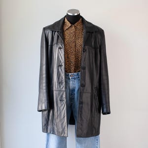 May include: A black leather coat with a button-up front, displayed on a mannequin. The coat is paired with a leopard print button-down shirt and light blue jeans. The coat has a classic, longline design.