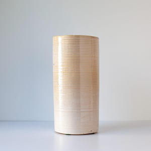May include: A tall, cylindrical vase with a textured, horizontal ribbed design. The vase has a light beige color with subtle variations in tone, creating a warm, neutral aesthetic. The vase is likely ceramic or stoneware.