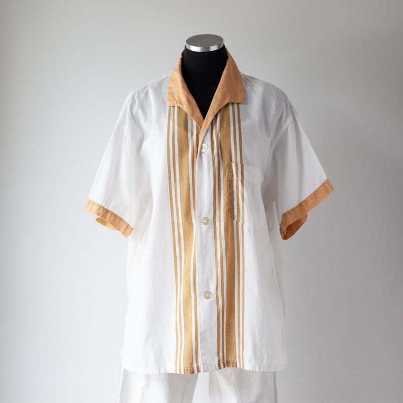 May include: White short-sleeved shirt with tan and white vertical stripes. The shirt has a tan collar and sleeve trim, and a single pocket. The shirt is button-down and appears to be made of a lightweight fabric.