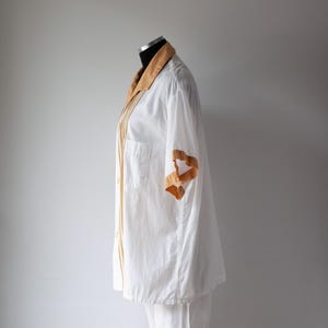 May include: White button-down shirt with a tan collar and sleeve accents. The shirt has a chest pocket and vertical tan stripes down the front. The sleeve has a tan ribbon design.