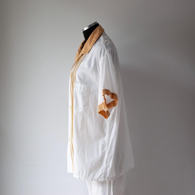 May include: White button-down shirt with a tan collar and sleeve accents. The shirt has a chest pocket and vertical tan stripes down the front. The sleeve has a tan ribbon design.