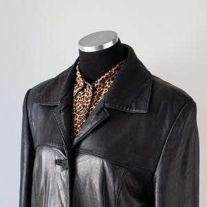 May include: A black leather jacket with a collared neckline and button closure. The jacket is worn over a button-down shirt with a brown and tan leopard print pattern. The jacket is displayed on a black mannequin.