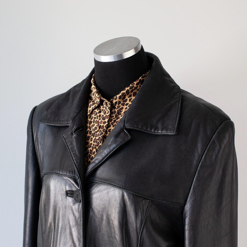 May include: A black leather jacket with a collared neckline and button closure. The jacket is worn over a button-down shirt with a brown and tan leopard print pattern. The jacket is displayed on a black mannequin.