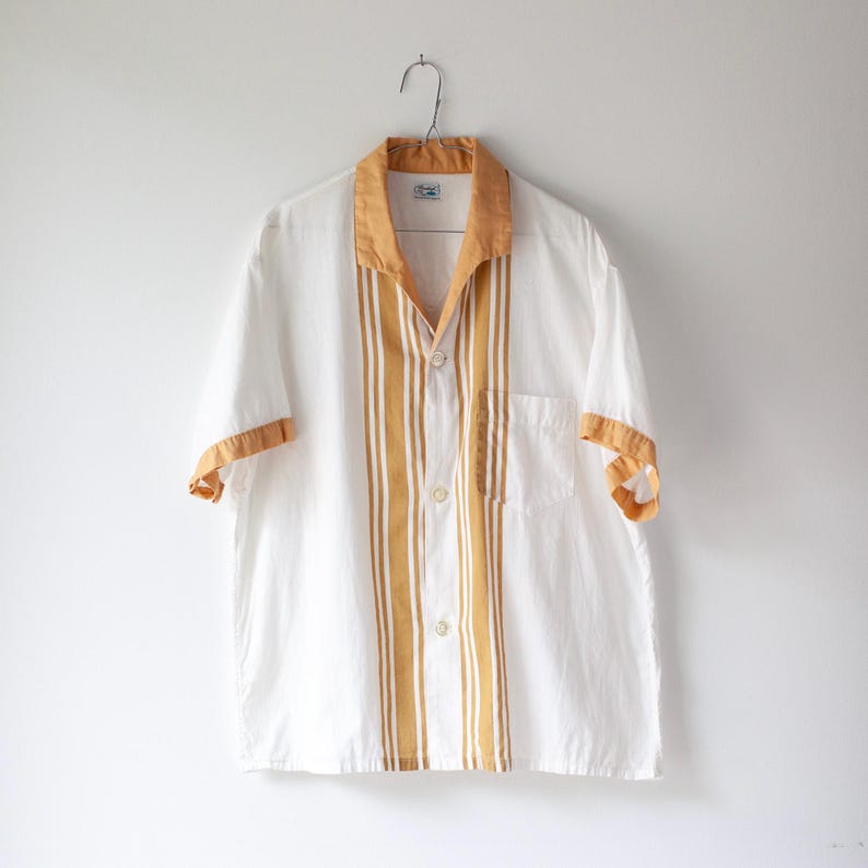May include: White short-sleeve shirt with a mustard yellow collar, cuffs, and vertical stripes down the front. The shirt has a button-down closure and a chest pocket. The shirt is hanging on a metal hanger.