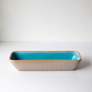 May include: Rectangular ceramic dish with a turquoise interior and a textured, beige exterior. The dish has rounded corners and a simple, modern design. The turquoise interior provides a striking contrast to the neutral exterior.
