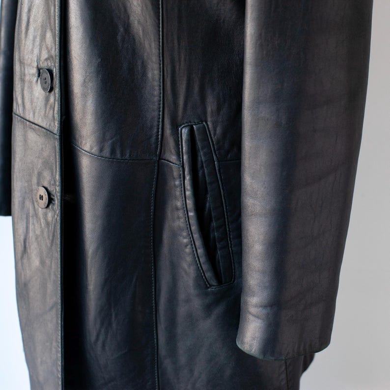 May include: A close-up of a navy blue leather coat. The coat features a button-down front, a pocket, and a long sleeve. The leather has a subtle sheen. The coat appears to be in good condition.