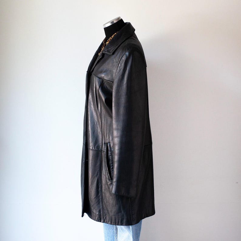 May include: A black leather coat with a collared neckline and a button-down front. The coat has two front pockets and a long, knee-length design. A gold chain necklace is visible at the neckline. The coat is displayed on a mannequin.