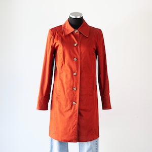 May include: A rust-colored, long-sleeved coat with a button-down front and a collared neckline. The coat is displayed on a mannequin and features several buttons down the front. The coat is approximately 40 inches long.