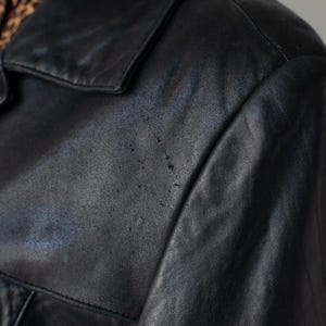 May include: Close-up of a black leather jacket. The jacket has a classic collar and a slightly worn appearance, with visible wear and tear on the leather. The jacket's texture is visible, showing the natural grain of the leather.