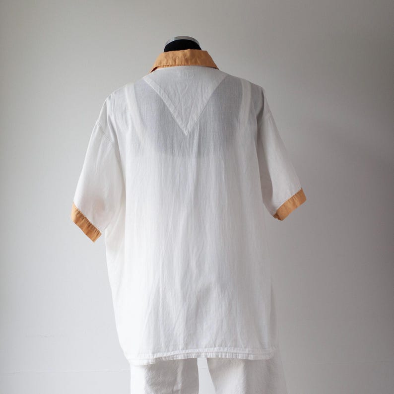 May include: White short-sleeved shirt with a tan collar and sleeve trim. The shirt is made of a lightweight fabric and has a relaxed fit. The shirt is displayed on a mannequin.
