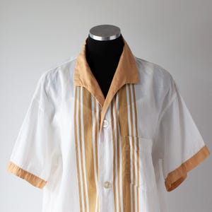 May include: White short-sleeve shirt with tan accents. The shirt features tan stripes down the front, a tan collar, and tan trim on the sleeves. A single pocket is visible on the chest.