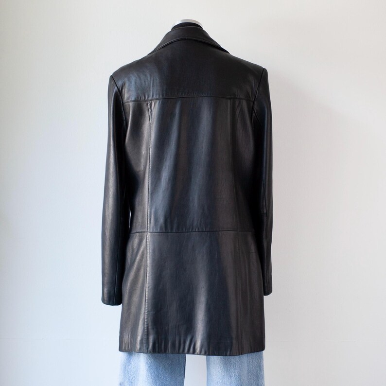 May include: A black leather coat with a notched collar and long sleeves. The coat is displayed on a mannequin against a white background. The coat has a tailored fit and a mid-thigh length.