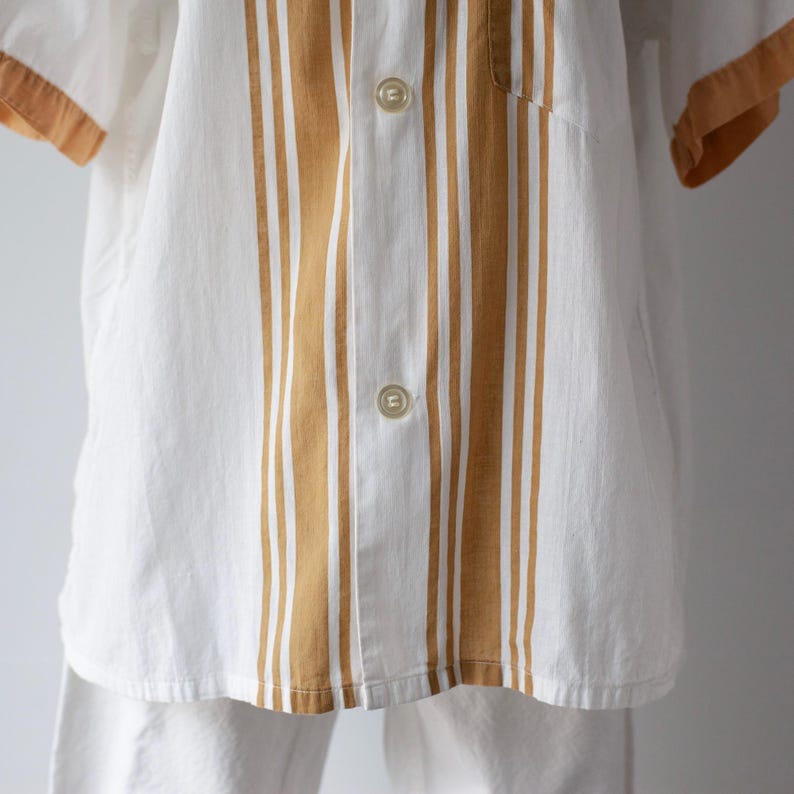 May include: White button-up shirt with vertical tan stripes down the center and tan trim on the short sleeves. The shirt has two visible buttons and is paired with white pants. The shirt is made of a lightweight fabric.