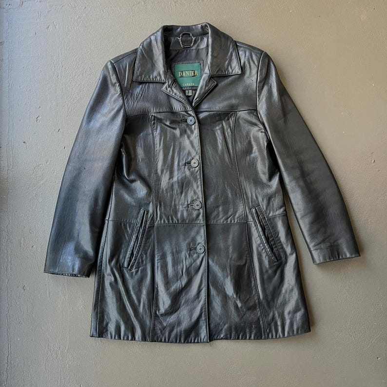 May include: A dark gray leather jacket with a collared neckline and button-up closure. The jacket has two front pockets and a label that reads "DANIER". The jacket is a mid-length coat.