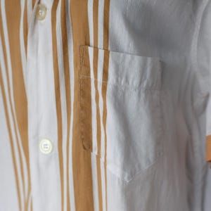 May include: Close-up of a white shirt with vertical brown stripes. The shirt has a pocket on the chest and a button-down collar. The sleeve cuff is a light orange color.