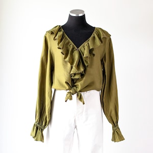 May include: An olive green long-sleeve blouse featuring a deep V-neckline with cascading ruffles, long sleeves with gathered ruffled cuffs, and a front tie-knot detail at the waist, displayed on a black mannequin torso over white pants.