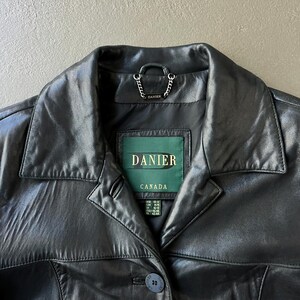 May include: Black leather jacket with a notched collar and button closure. The jacket features a green label with the brand name "DANIER" and "CANADA" printed on it. A small chain detail is visible near the collar.