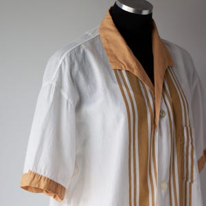 May include: White short-sleeved shirt with a tan collar, sleeve cuffs, and vertical stripes down the front. The shirt has a button-down closure and a chest pocket. The shirt is displayed on a mannequin.