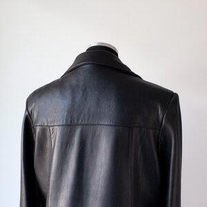 May include: A black leather jacket displayed on a mannequin. The jacket features a classic collar and a tailored fit. The back of the jacket is smooth and the leather appears to be in good condition. The background is a plain white wall.