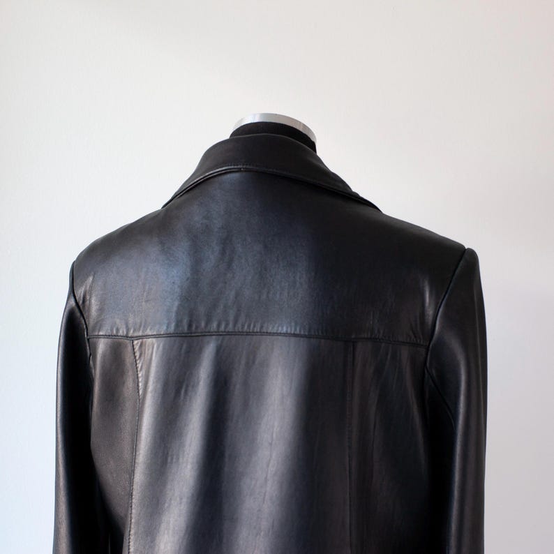 May include: A black leather jacket displayed on a mannequin. The jacket features a classic collar and a tailored fit. The back of the jacket is smooth and the leather appears to be in good condition. The background is a plain white wall.