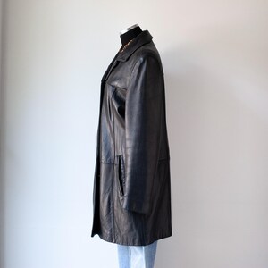 May include: A black leather coat displayed on a mannequin. The coat has a classic design with a collared neckline, front pockets, and a button-down closure. The coat is knee-length and appears to be in good condition. The mannequin is wearing blue jeans.