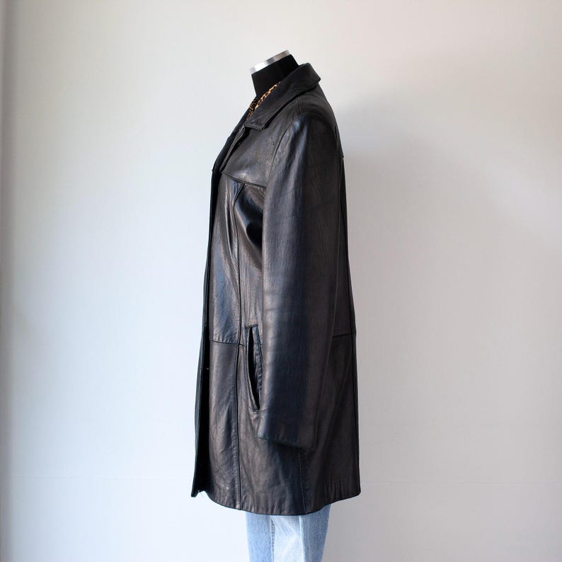 May include: A black leather coat displayed on a mannequin. The coat has a classic design with a collared neckline, front pockets, and a button-down closure. The coat is knee-length and appears to be in good condition. The mannequin is wearing blue jeans.