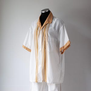 May include: White short-sleeved shirt with tan accents on the collar and sleeves. Vertical tan stripes run down the front of the shirt. The shirt is displayed on a mannequin. The shirt has a pocket on the left side.