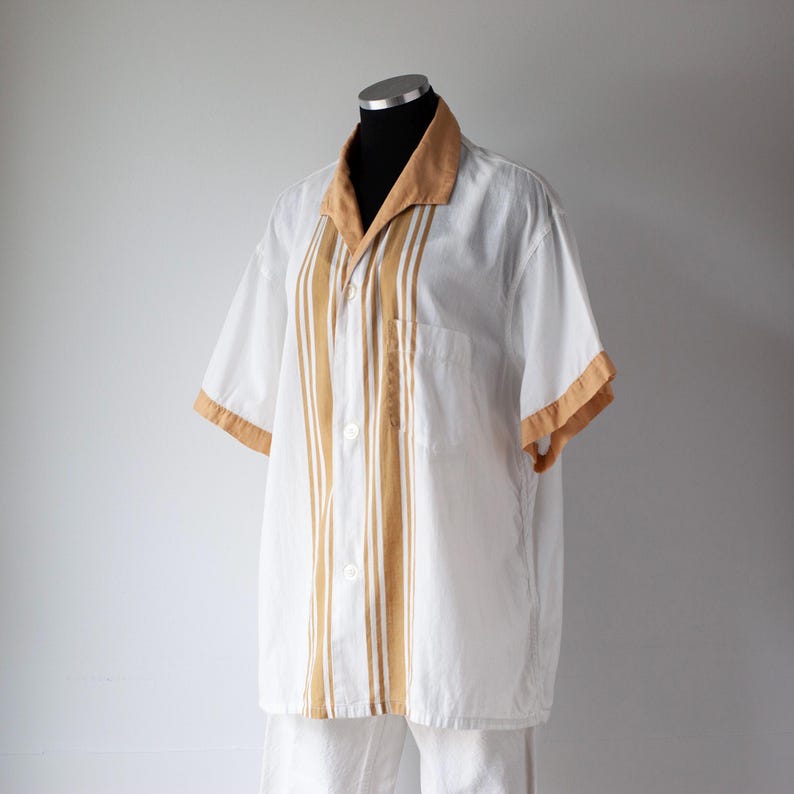 May include: White short-sleeved shirt with tan accents on the collar and sleeves. Vertical tan stripes run down the front of the shirt. The shirt is displayed on a mannequin. The shirt has a pocket on the left side.