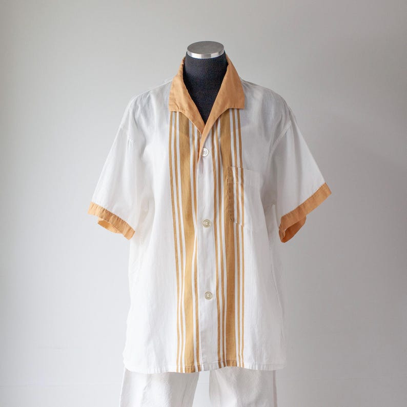 May include: White short-sleeved shirt with tan accents. The shirt features tan stripes down the front and on the sleeves, with a matching collar. The shirt is button-down and has a pocket on the left side.
