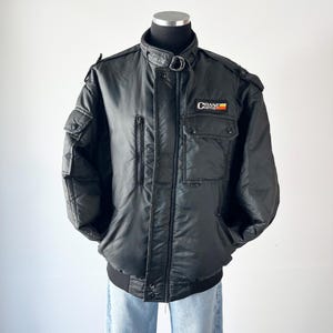 May include: Black bomber jacket with multiple pockets, a zip closure, and a stand-up collar. The jacket features the "CRANE Cams" logo on the chest. The jacket is displayed on a mannequin.