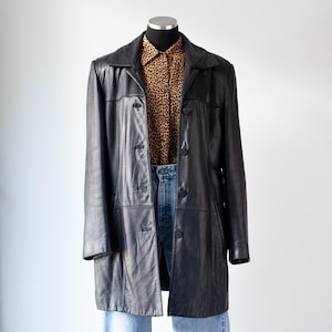 May include: A black leather coat with a button-up front, displayed on a mannequin. The coat is paired with a leopard print button-down shirt and blue jeans. The coat has a classic, longline silhouette.