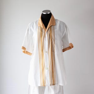 May include: White short-sleeved shirt with tan accents. The shirt features a tan collar, sleeve cuffs, and vertical stripes down the front. It has a button-down closure and a chest pocket. The shirt is displayed on a mannequin.