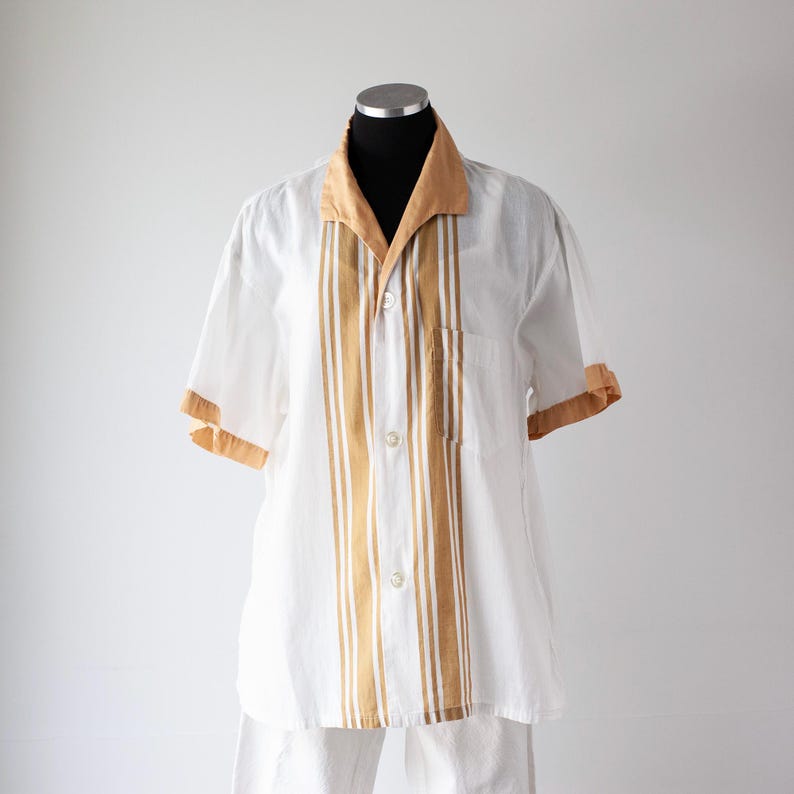 May include: White short-sleeved shirt with tan accents. The shirt features a tan collar, sleeve cuffs, and vertical stripes down the front. It has a button-down closure and a chest pocket. The shirt is displayed on a mannequin.