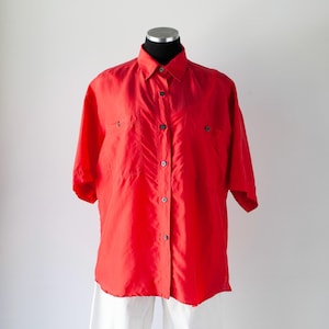 May include: A vibrant red short-sleeved button-down shirt featuring a classic collar, a central vertical row of dark buttons, and two chest pockets. The garment is displayed on a mannequin, showcasing a smooth, lightweight fabric texture.