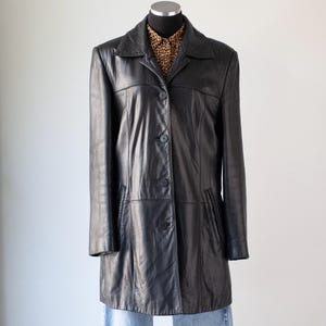May include: A black leather coat with a collared neckline and button-down front. The coat has a tailored fit and features pockets. A leopard print shirt is visible underneath the coat.