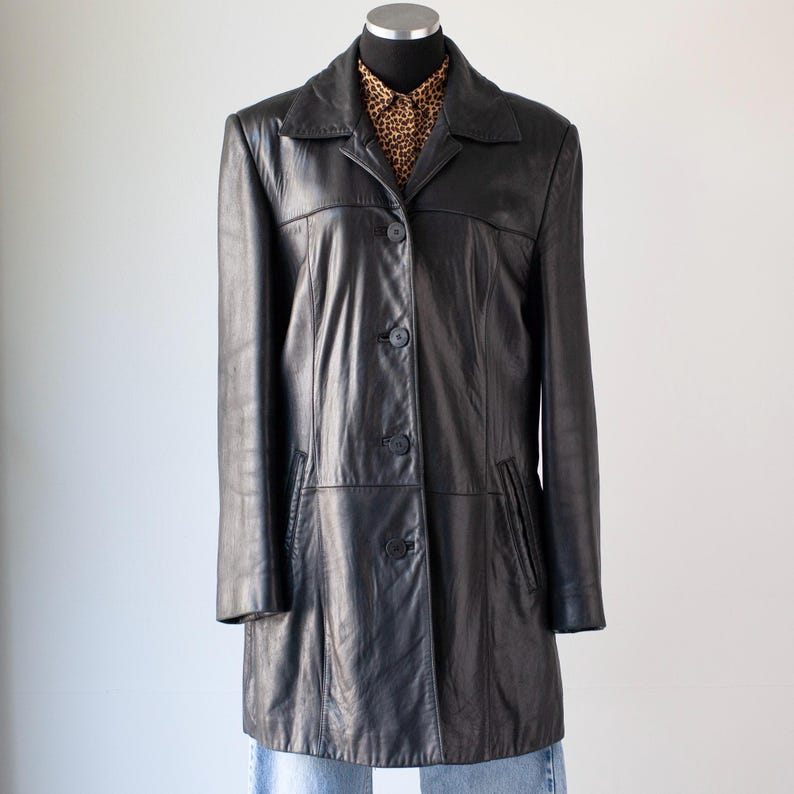May include: A black leather coat with a collared neckline and button-down front. The coat has a tailored fit and features pockets. A leopard print shirt is visible underneath the coat.