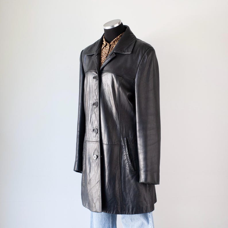 May include: A black leather coat with a collared neckline and button-down closure. The coat has long sleeves and a mid-length cut, displayed on a mannequin. A patterned shirt is visible underneath the coat.