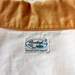 May include: Close-up of a vintage shirt collar with a mustard yellow collar and a white body. The shirt tag reads "Pennleigh Wash and Wear Deluxe" with the text "100% Cotton W.T. Grant Co."