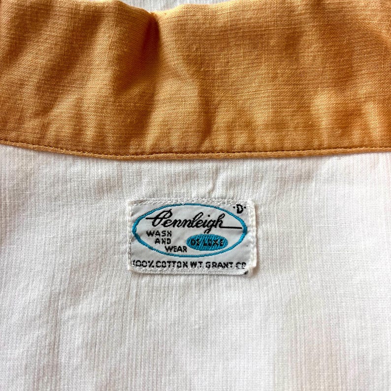 May include: Close-up of a vintage shirt collar with a mustard yellow collar and a white body. The shirt tag reads "Pennleigh Wash and Wear Deluxe" with the text "100% Cotton W.T. Grant Co."